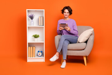 Full size portrait of charming person sitting chair use tablet typing email isolated on orange color background