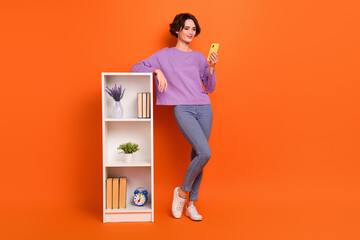 Full size photo of pretty nice lady use gadget telephone stand near book shelf isolated on orange...