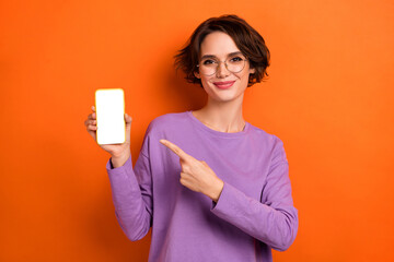 Photo of charming pretty girl dressed purple pullover eyewear pointing modern gadget empty space isolated orange color background