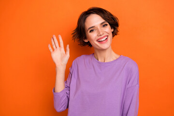 Photo of excited adorable lady wear violet sweatshirt waving arm hello smiling isolated orange color background