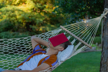 Young man resting and sleeping in a hammock in spring time with a red book covering his eyes and face