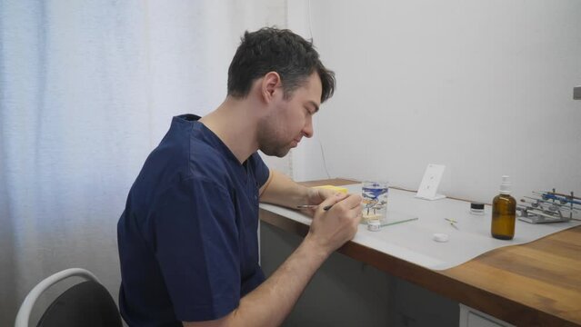 Stomatology technician deals with the dental crown production. Laboratory worker produces metal dental crowns for dentistry. Dental technician painting the dental crown product using a brush tool
