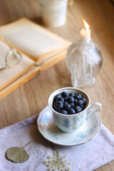 Vintage porcelain cup filled with fresh blueberries, pressed flowers, open book, reading glasses and lit candle on the table. Hygge at home. Selective focus.