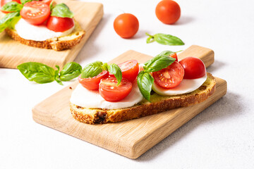 Fresh tasty caprese sandwiches with full grain bread, mozzarella cheese, basil and cherry tomatoes on the serving board on white stone table.