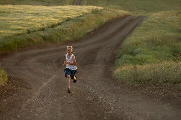 Obraz premium A boy in white t-shirt runs at sunset along a rural road next to a wheat field. Summer holidays in the countryside. Happy childhood