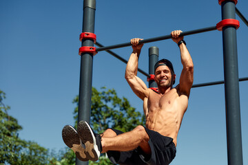 Athletic young man hanging from the bars at the calisthenics gym outdoors smiling