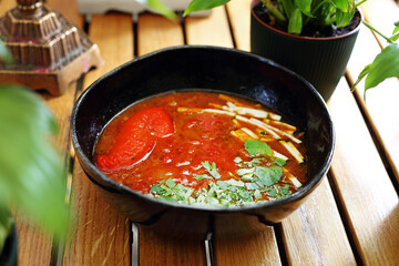 Tomato soup with fresh herbs in a modern black bowl, on a wooden table, horizontal view, close-up.