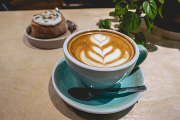 Colorful cup with latte coffee with flower design, delicious cinnamon roll cake and green decorative plant in a coffee shop interior
