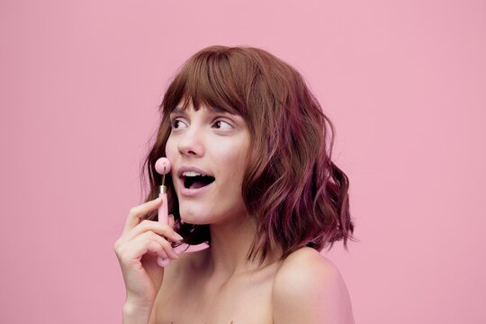 A Happy, Enthusiastic Woman, Standing On A Pink Background With Pink Lighting From The Side, With Her Hair Styled, Doing A Facial Massage With A Pink Roller, Her Mouth Wide Open In Surprise.