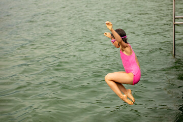 Little sporty girl jumping into water of lake from wooden pier on cloudy summer day