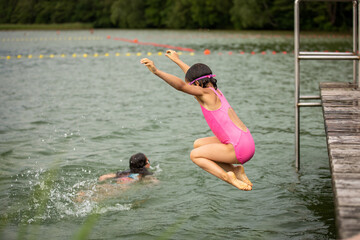 Little sporty girl jumping into water of lake from wooden pier on cloudy summer day