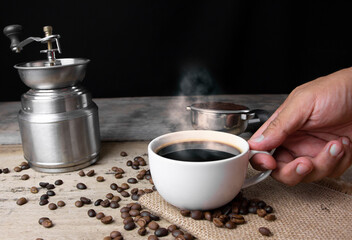 Front view hand holding cup of black Americano coffee on a wooden table with a pile of roasted Arabica coffee beans on black background.