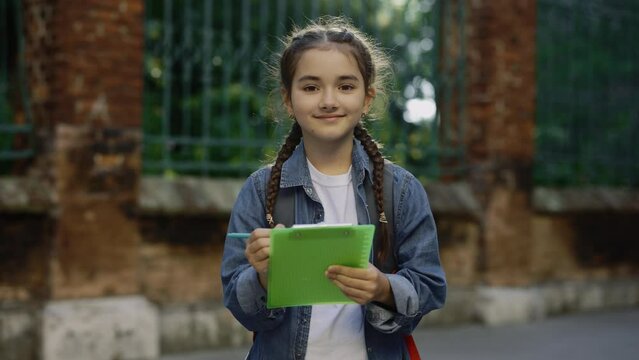 Portrait of lillte cute casual girl braids with schoolbag standing schoolyard smilling and writting notes outside. Education concept