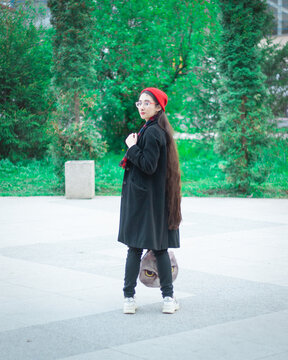 A Young Woman With Long Hair And A Red Hat Wearing Glasses And Dark Coat With A Red Scarf In The Park Looking Backwards Against Trees On A Pedestrian Road.