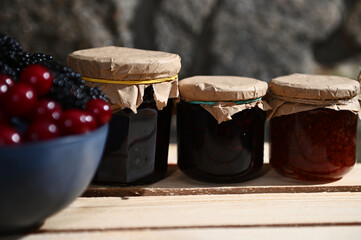 Assorted berry jam in jars on a wooden crate. Partial view of blurred bowl with fresh ripe berries on the foreground