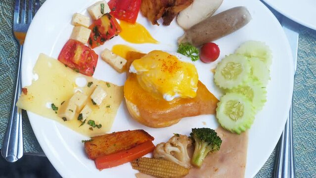 Benedict egg with smoked salmon and hollandaise sauce, fork and knife. Plate with poached egg, bacon, sausage, cheese, grilled vegetables on buffet breakfast, brunch in restaurant at hotel, resort.