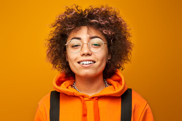 Positive kazakh student girl with afro hairstyle and backpack smiling on yellow studio background