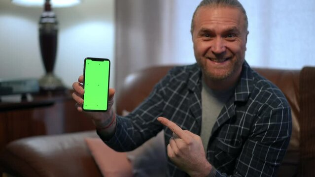 Happy satisfied man showing green screen phone pointing smiling looking at camera. Portrait of Caucasian gamer posing winning in online betting on smartphone chromakey app mockup
