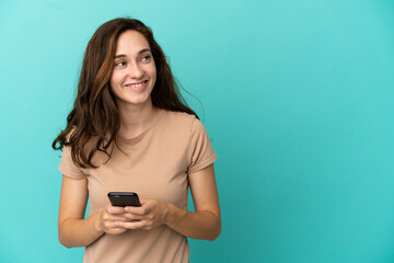 Young caucasian woman isolated on blue background using mobile phone and looking up