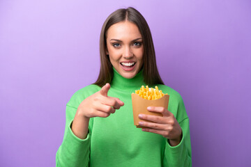 Young caucasian woman holding fried chips on purple background surprised and pointing front