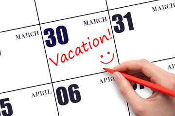 A hand writing a VACATION text and drawing a smiling face on a calendar date 30 March. Vacation...