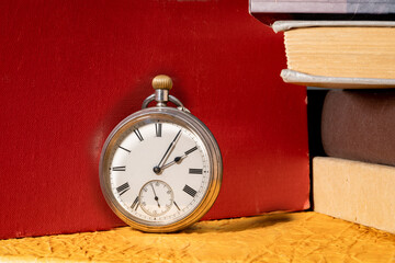 Antique silver retro clock on background of stacks of books in library. Round old vintage clock with dial and hands near books with red, yellow and gray covers.