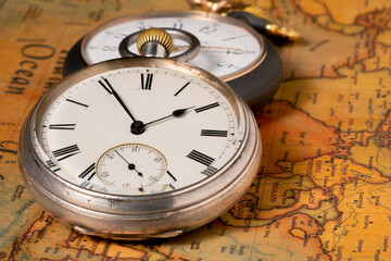 Two silver retro pocket watches lying on an old paper map of the world. Antique gray round clock with a dial and hands on a geographical map with continents and countries.