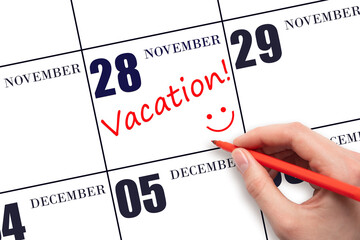 A hand writing a VACATION text and drawing a smiling face on a calendar date 28 November. Vacation planning concept.