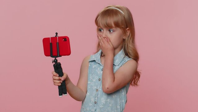 Wow reaction. Impressed young preteen child girl kid blogger taking selfie on mobile phone selfie stick, communicating video call online. Little toddler children isolated on studio pink background