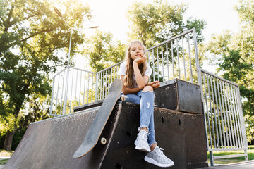 Child girl sitting with skate board on sport ramp. Sports equipment for kids. Active teenager with skate board on skate park playground.