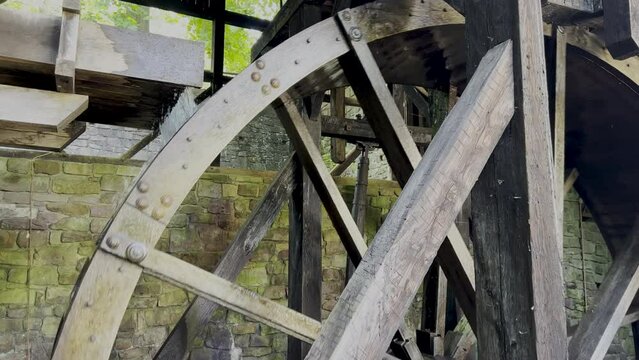 4K footage of a rustic turning colonial water wheel in a stone structure with upper portion open to green nature. Wheel is very large and the wood is dark and rustic, water pours from sluice