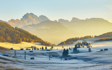 Beautiful view of traditional wooden mountain chalets on scenic Alpe di Siusi with famous Langkofel mountain peaks in the background in golden morning light at sunrise, Dolomites, Italy