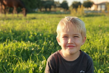 A smiling 4-year-old little blond toddler boy is sitting in green yellow sunny autumn meadow and smiling