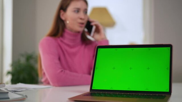 Green screen of laptop with blurred woman talking on phone at background. Busy Caucasian businesswoman freelancer discussing e-business profitability with chromakey web page template at front
