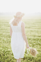 Fototapeta na wymiar A beautiful young girl in a white dress and a wreath on her head walks through the field with a bouquet of wheat ears. The concept of ecology and nature. Green field in the rays of the morning sun