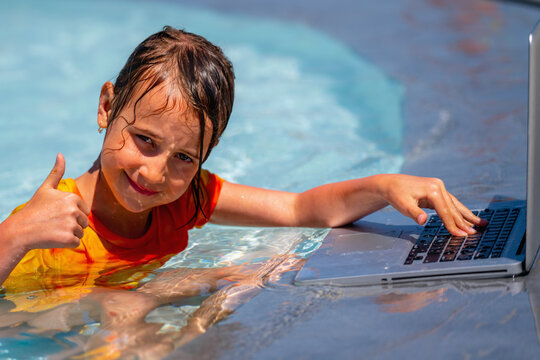 Beautiful young girl gesturing thumb up, using laptop and working online remotely in the swimming pool. Summer vacations, rest and work at the same time. Horizontal image.