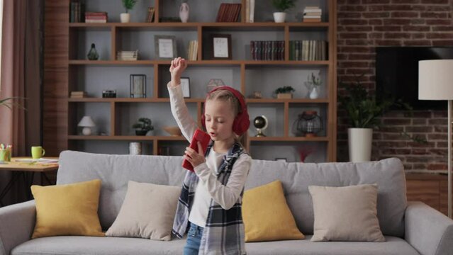 Happy little girl wearing red headphones dancing and singing using smartphone like microphone. Cute child enjoy listening favorite music tracks in mobile application Home leisure, happy childhood.