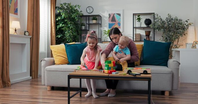 A woman sits watching two children at home. Caregiver entertains toddlers with toys, shows blocks to youngest boy, girl in pink dress plays dulcimer, babysitting.