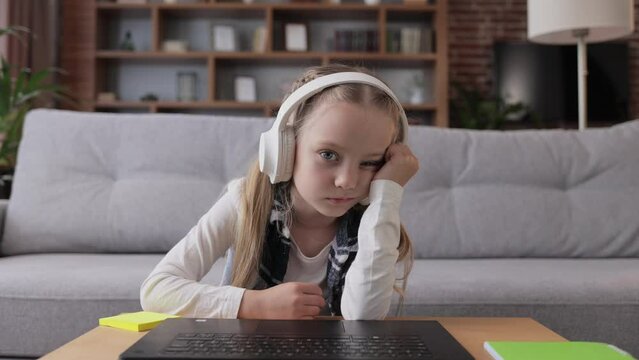 Cute child studying remotely at home using laptop. Tired primary schoolgirl falling asleep during an online lesson with teacher. Distance learning is boring and uninteresting. Web cam view.