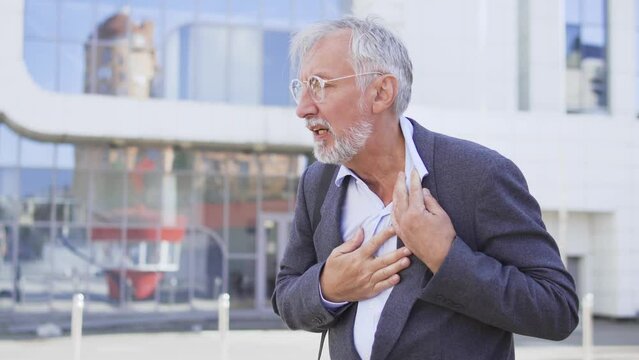 Mature man in eyeglasses feeling hardness of breathe and pain in heart, unbuttoning shirt to breathe deeply, risk of heart attack, emergency situation outdoor, call 911