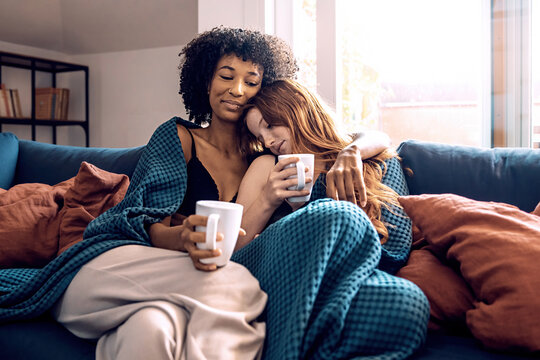 Multiethnic lesbian couple hugging and drinking coffee on sofa at home - LGBT lifestyle concept