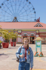 Obraz na płótnie Canvas A cheerful woman with pigtails in a denim outfit walks in a sunny amusement park against the backdrop of a ferris wheel. Weekend for a walk