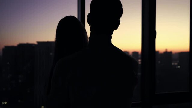 Silhouette of a guy hugging a girl against the backdrop of panoramic windows overlooking the city and sunset. The camera zooms in on the couple from behind