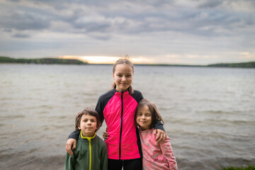 Fototapeta na wymiar Three smiling kids standing against lake on summer cloudy day