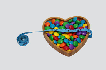Heart-shaped bowl with colorful candies and a measuring tape on a white background, top view,...