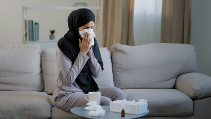 Sick arabian muslim young unhealthy woman in hijab suffer from runny nose flu disease seasonal allergy sneeze blow nose in paper napkin symptoms of coronavirus drink hot tea sit at home self-isolation