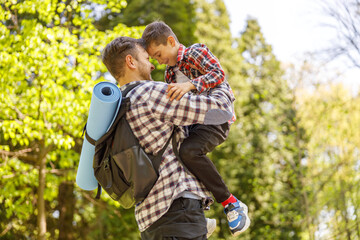 Cheerful Caucasian father with backpack holding small son on hands in park on summer day.