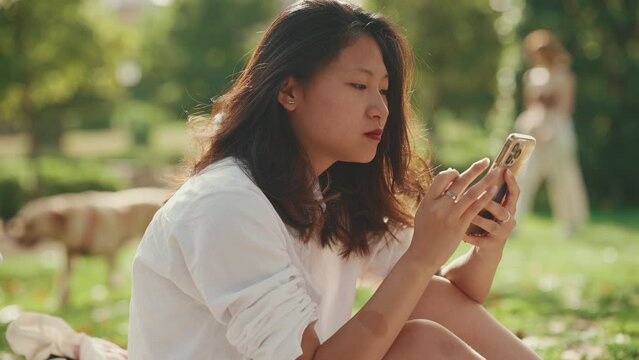 Young Asian woman with long brown hair wearing white t-shirt sitting in park having picnic on summer day outdoors, using cell phone. Girl browsing social networks, photo, video on smartphone