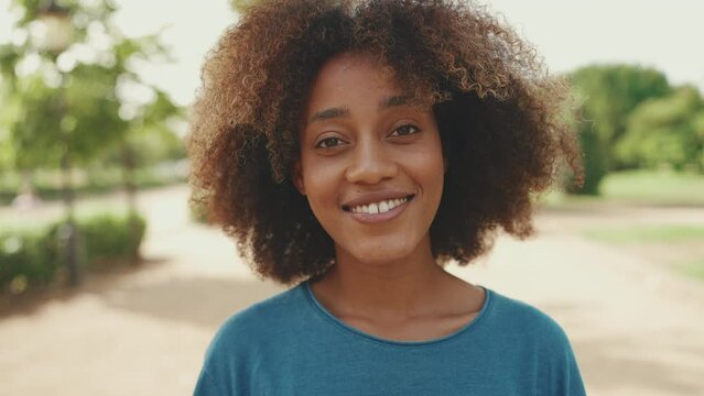 Close-up of young smiling woman with curly hair wearing blue t-shirt posing for the camera in the park. The girl smiles and says hello to the camera