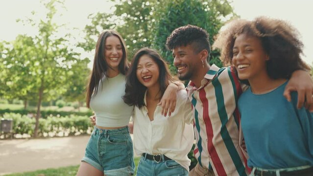 Happy multiethnic young people walk embracing on summer day outdoors. Group of friends are talking and laughing merrily while walking along path in city park